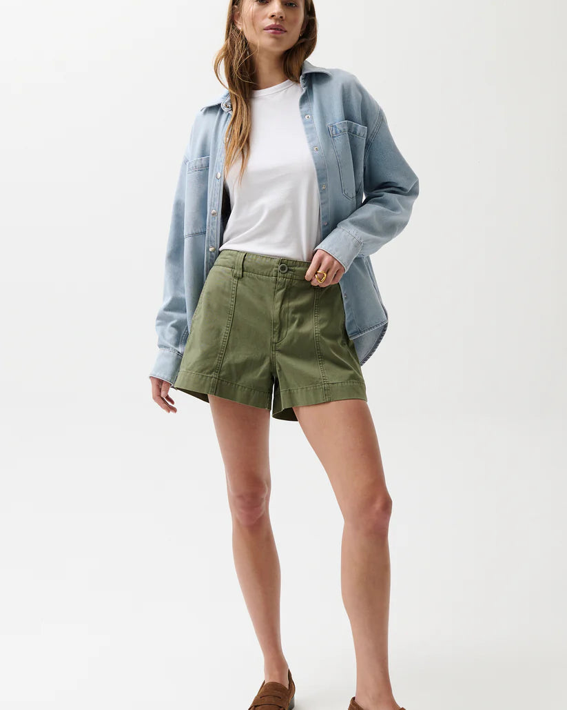 Woman wearing a light blue denim shirt, white t-shirt, green shorts, and brown loafers on a white background