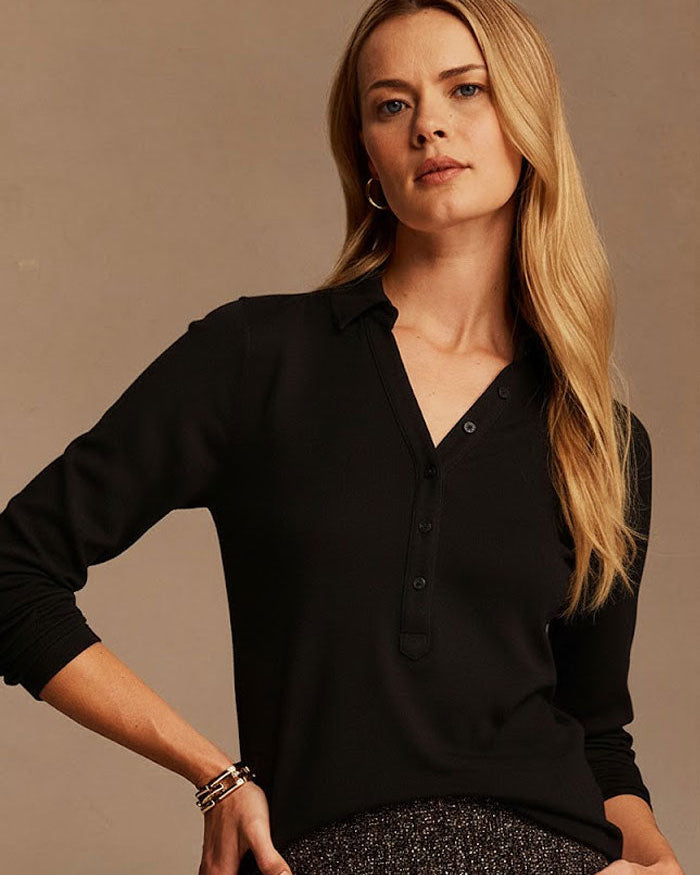A blonde woman wearing a black long-sleeve button-front collared top and a dark textured skirt, posing against a warm beige studio background.