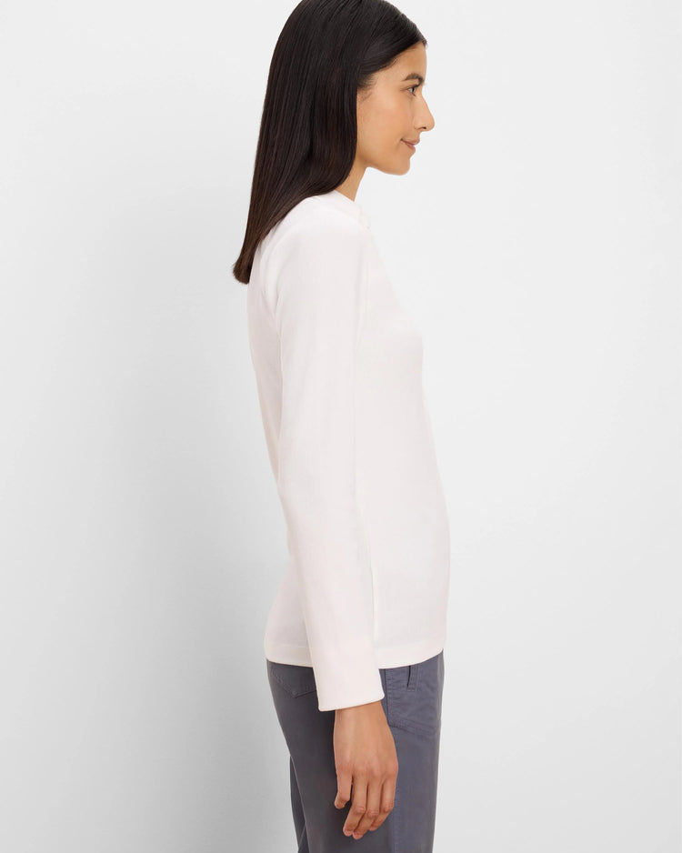 A woman shown in side profile wearing a light pink long-sleeve knit top and gray trousers against a plain light gray studio background.