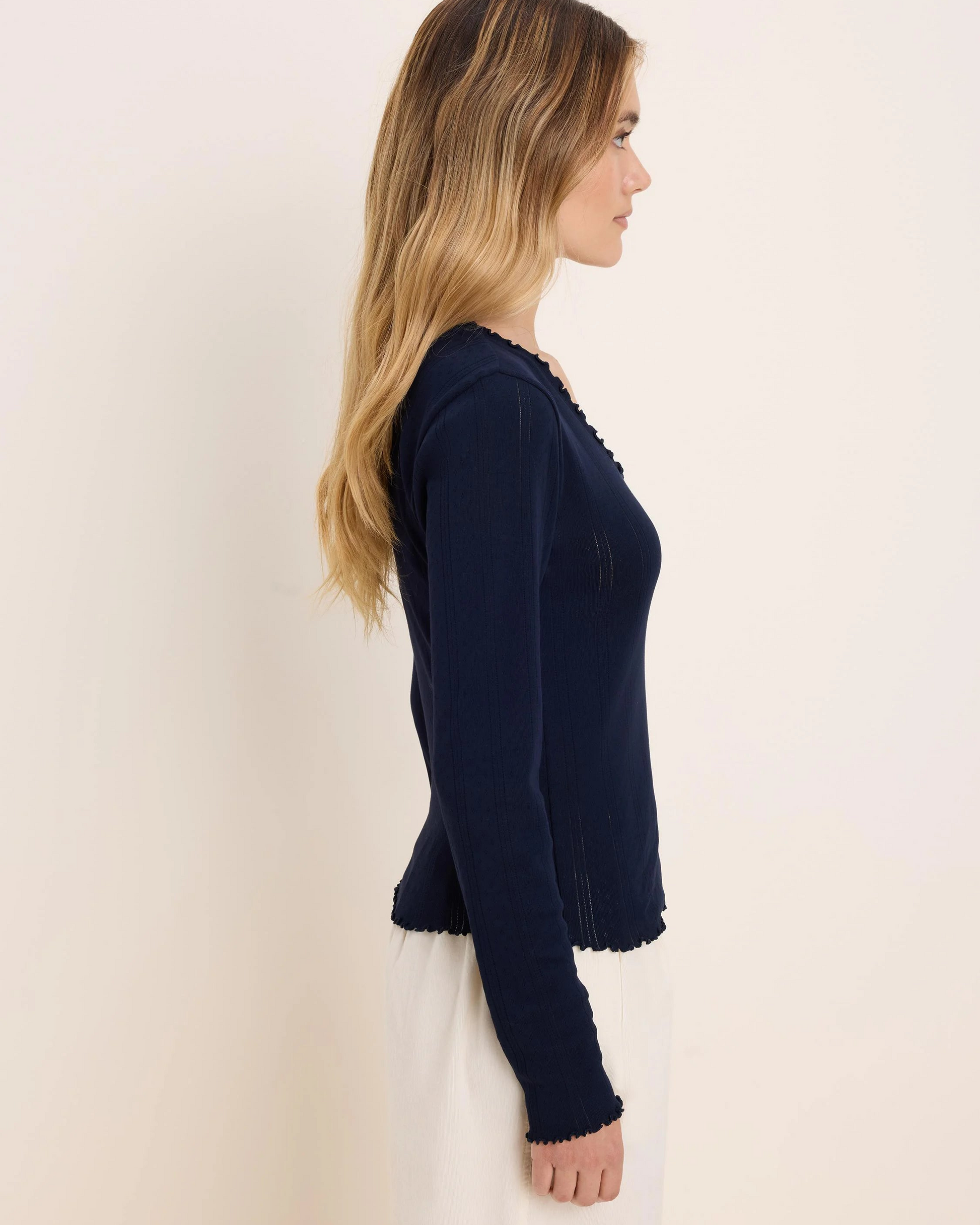A woman shown in side profile wearing a navy blue long-sleeve knit top with a scalloped neckline and hem, paired with a white skirt, against a plain off-white background.
