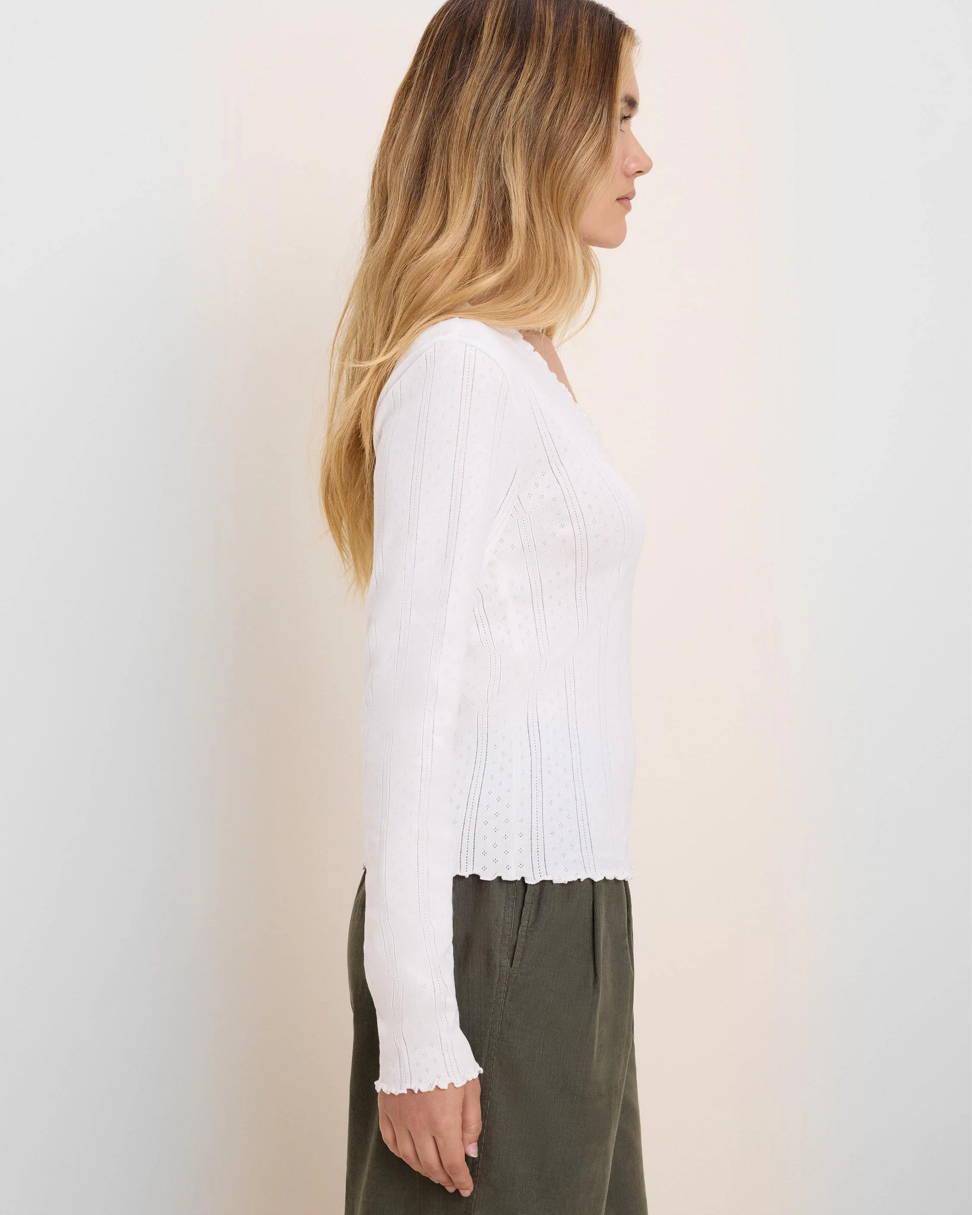A woman shown in side profile wearing a white long-sleeve knit top with a scalloped hem and olive green wide-leg pants, standing against a plain off-white wall.