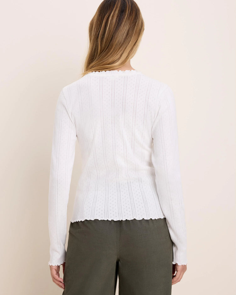 A woman shown from the back wearing a white ribbed knit long-sleeve top with a scalloped hem and olive green pants, against a plain beige studio background.