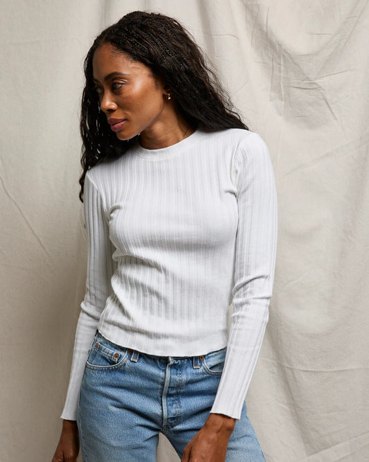 Woman wearing a white ribbed long-sleeve top and blue jeans against a neutral background