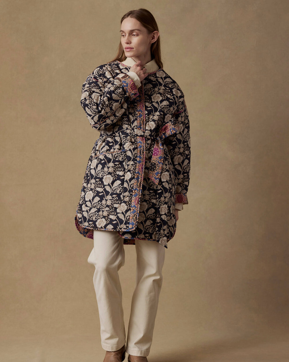 Person wearing a patterned coat and white pants against a beige background
