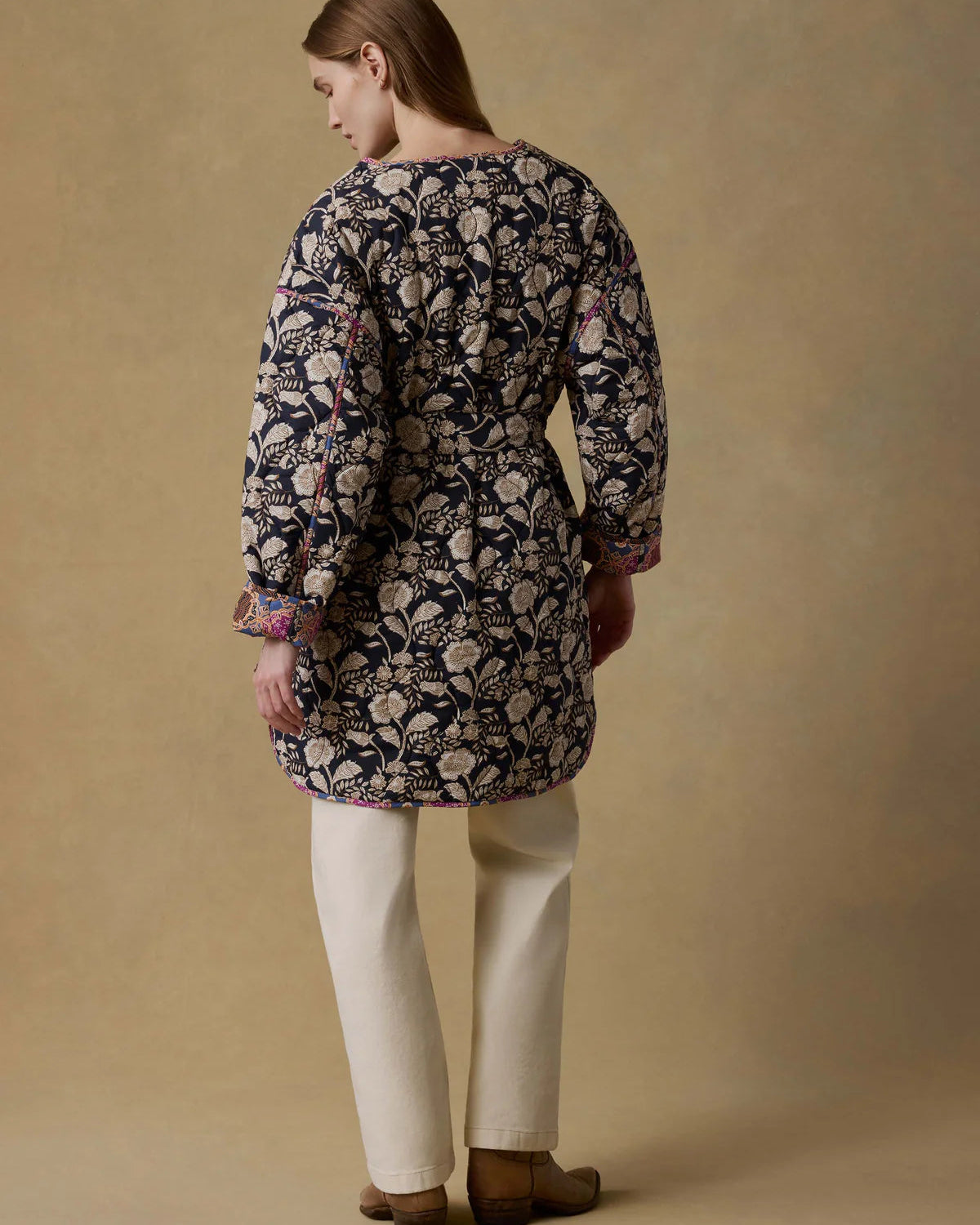 Person wearing a patterned long-sleeve top and white pants on a beige background