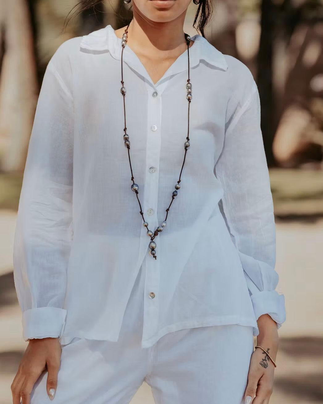 Person wearing a white shirt and light pants with a long necklace, standing outdoors.