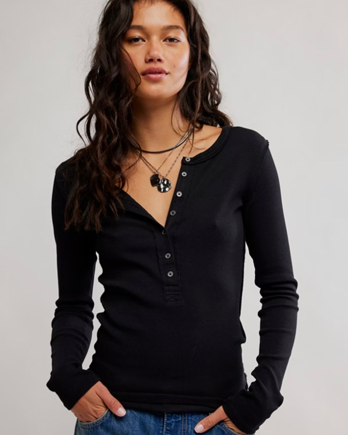 A woman wearing a black long-sleeve henley top with a button placket, layered with black necklaces, paired with blue jeans, against a plain light gray background.