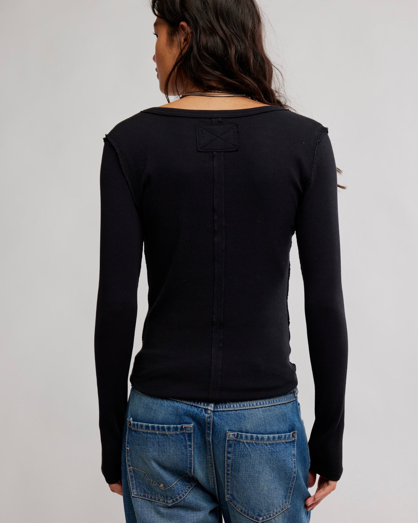 A woman shown from the back wearing a fitted black long-sleeve top with a small rectangular patch detail at the upper back, paired with blue jeans, against a plain light gray studio background.
