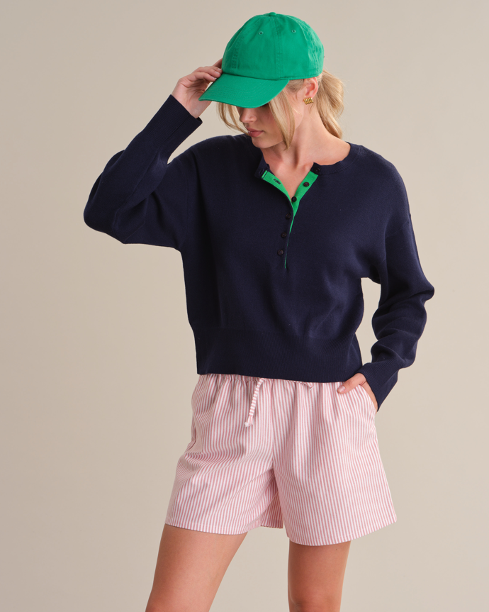 Person wearing a navy sweater, pink shorts, and green cap on a beige background