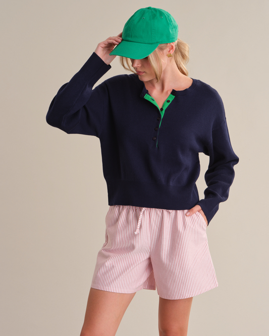 Person wearing a navy sweater, pink shorts, and green cap on a beige background