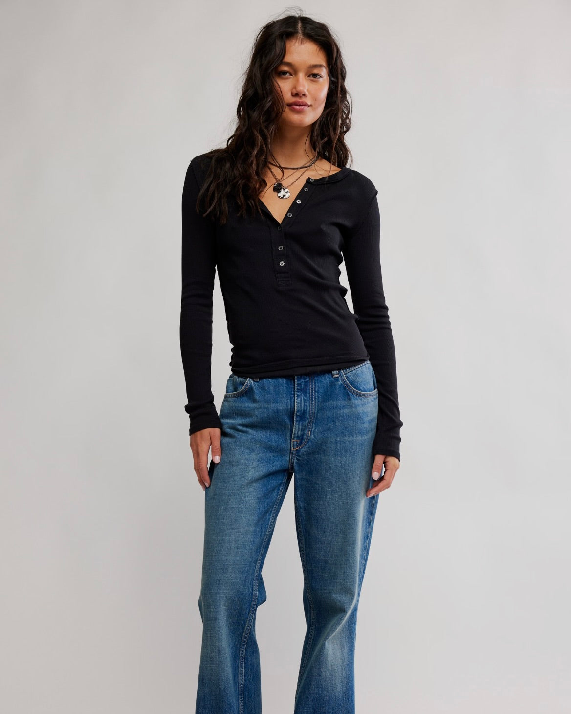 A woman modeling a black long-sleeve henley top with blue jeans against a plain light gray studio background.