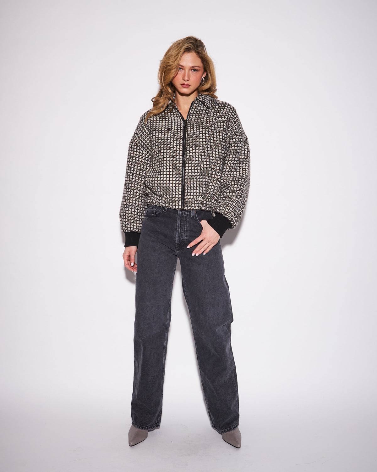 A woman stands against a plain light gray studio background wearing a black-and-white small-scale patterned zip-front jacket with dark charcoal wide-leg jeans and taupe pointed shoes.