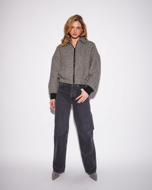A woman stands against a plain light gray studio background wearing a black-and-white small-scale patterned zip-front jacket with dark charcoal wide-leg jeans and taupe pointed shoes.