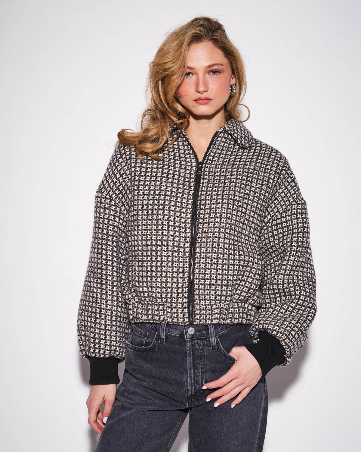 A woman modeling a cropped zip-front jacket with a black and beige geometric pattern, black ribbed cuffs, and dark gray jeans against a plain white studio background.