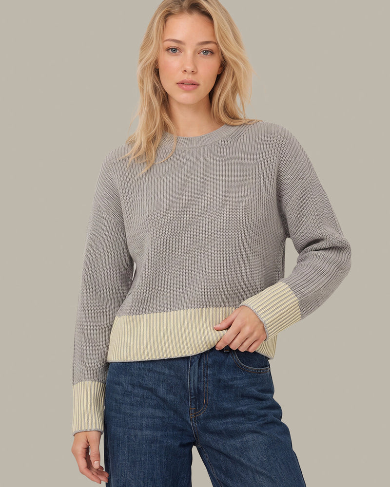 A woman modeling a ribbed knit sweater with a light gray body and contrasting cream ribbed cuffs and hem, paired with blue jeans against a solid light gray background.
