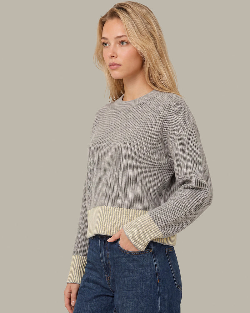 A woman wearing a ribbed knit sweater with a light gray body and cream-colored ribbed trim at the hem and cuffs, paired with blue jeans, against a solid light gray background.