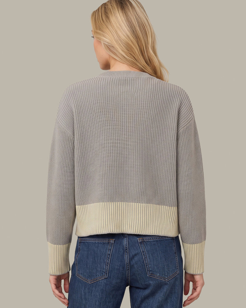 A woman shown from the back wearing a ribbed knit sweater with a light gray body and contrasting cream ribbed bands at the hem and cuffs, paired with blue jeans against a solid taupe background.
