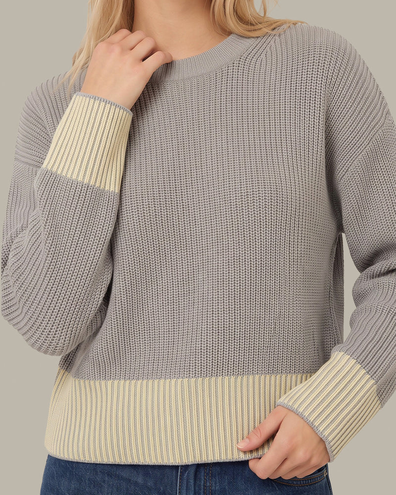 A woman wearing a gray ribbed knit sweater with contrasting cream ribbed cuffs and hem, paired with blue jeans against a neutral studio background.