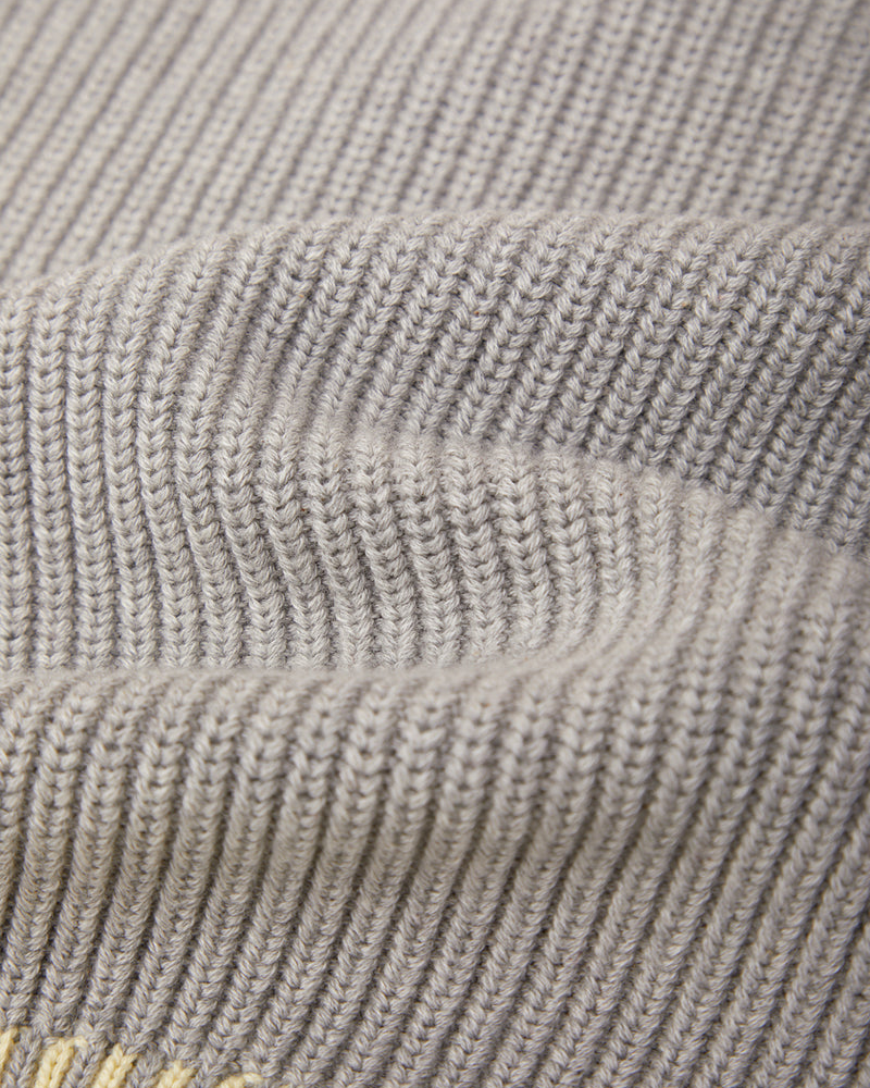 Close-up of a ribbed knit fabric in a light warm gray or taupe color, showing the texture and stitch pattern.