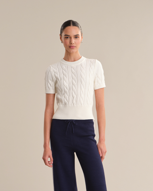 Woman wearing a white cable knit sweater and navy pants on a beige background