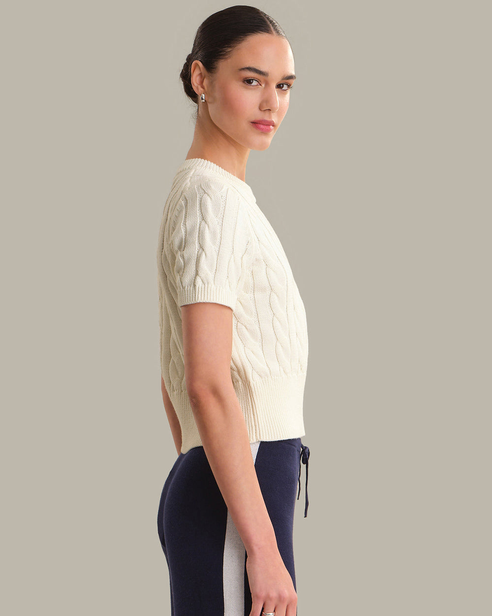 Woman wearing a cream cable knit sweater and dark pants against a gray background