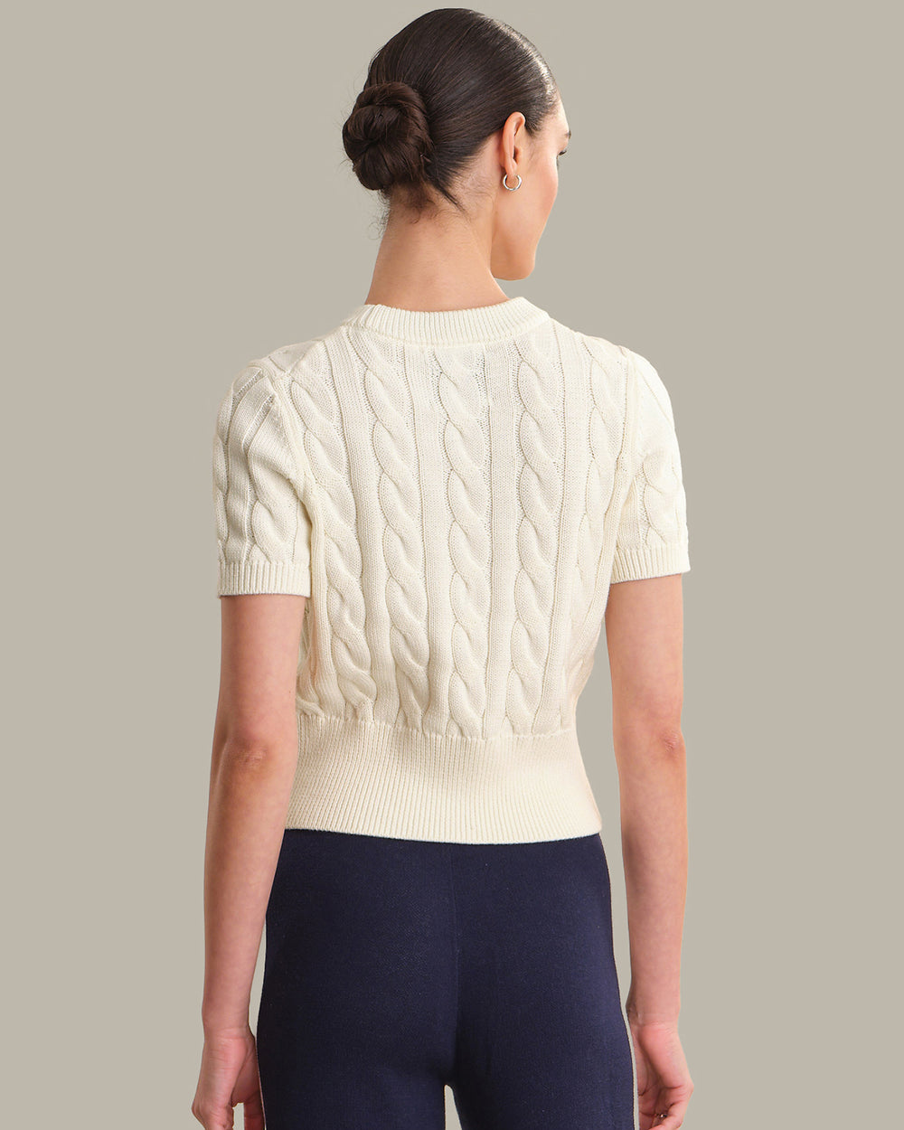 Woman wearing a cream cable knit sweater and navy pants against a gray background