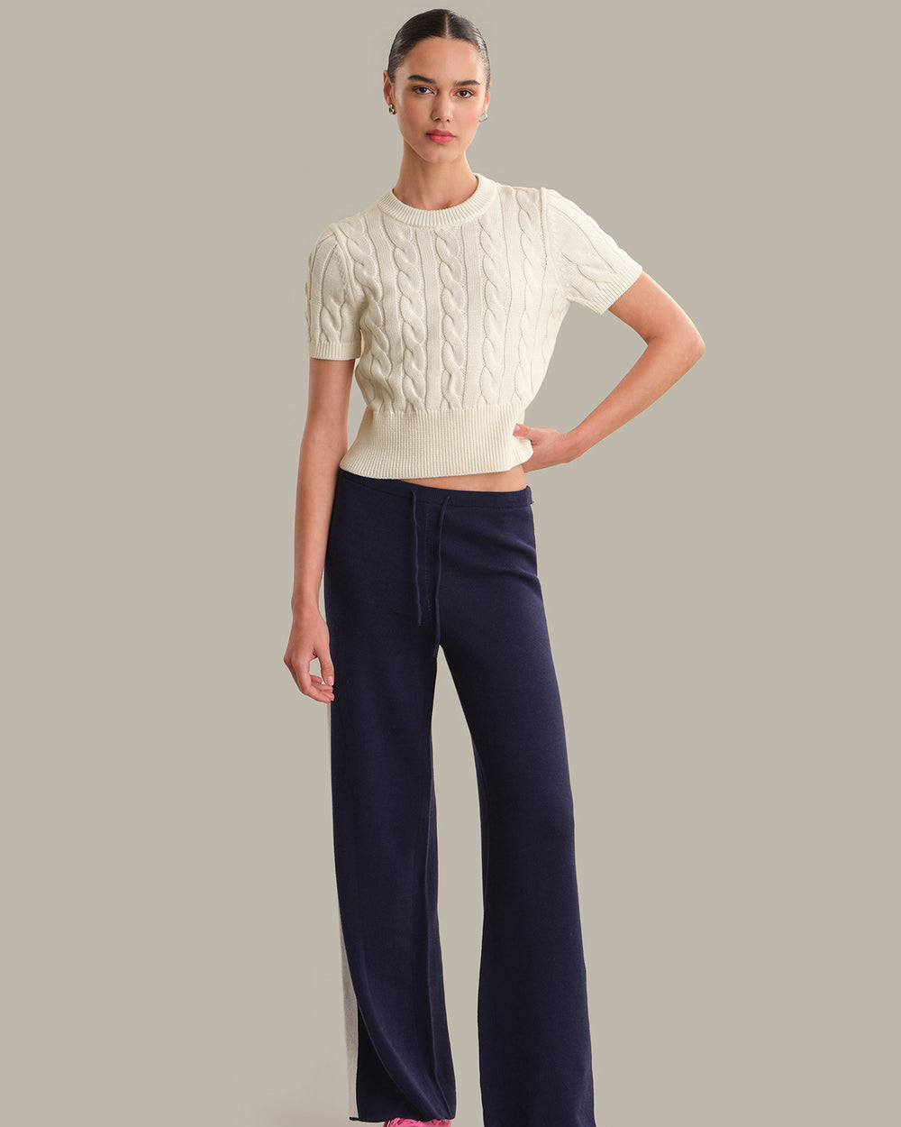 Woman wearing a white cable knit sweater and navy pants on a gray background