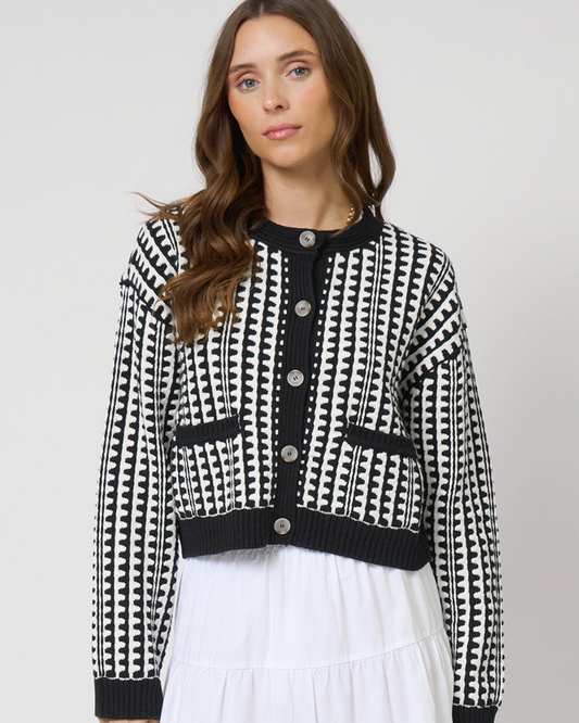 Woman wearing a black and white patterned cardigan over a white dress on a plain background