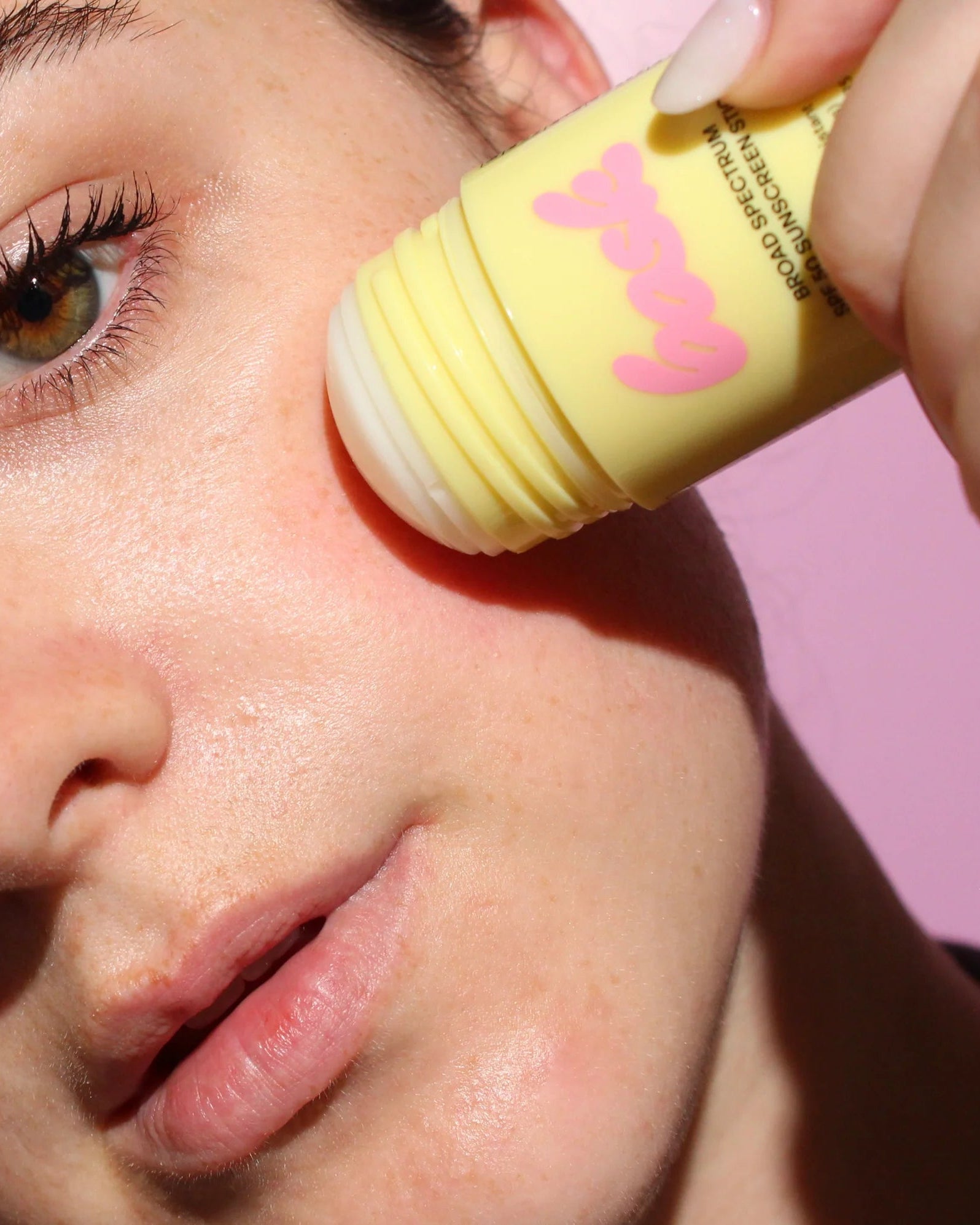 Person applying a yellow cosmetic product to their face with a pink applicator on a pink background