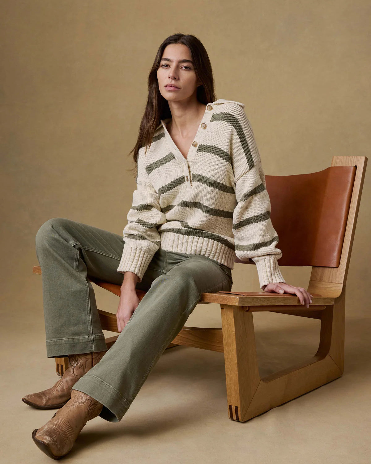 Woman sitting on a wooden chair wearing a striped sweater and green pants against a beige background
