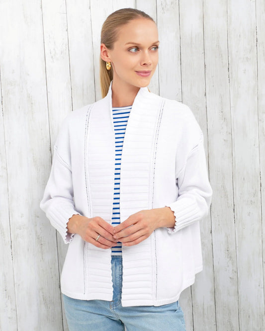 A woman wearing a white open-front cardigan with vertical pleating over a blue and white striped shirt and light blue jeans, standing against a white wood plank wall.