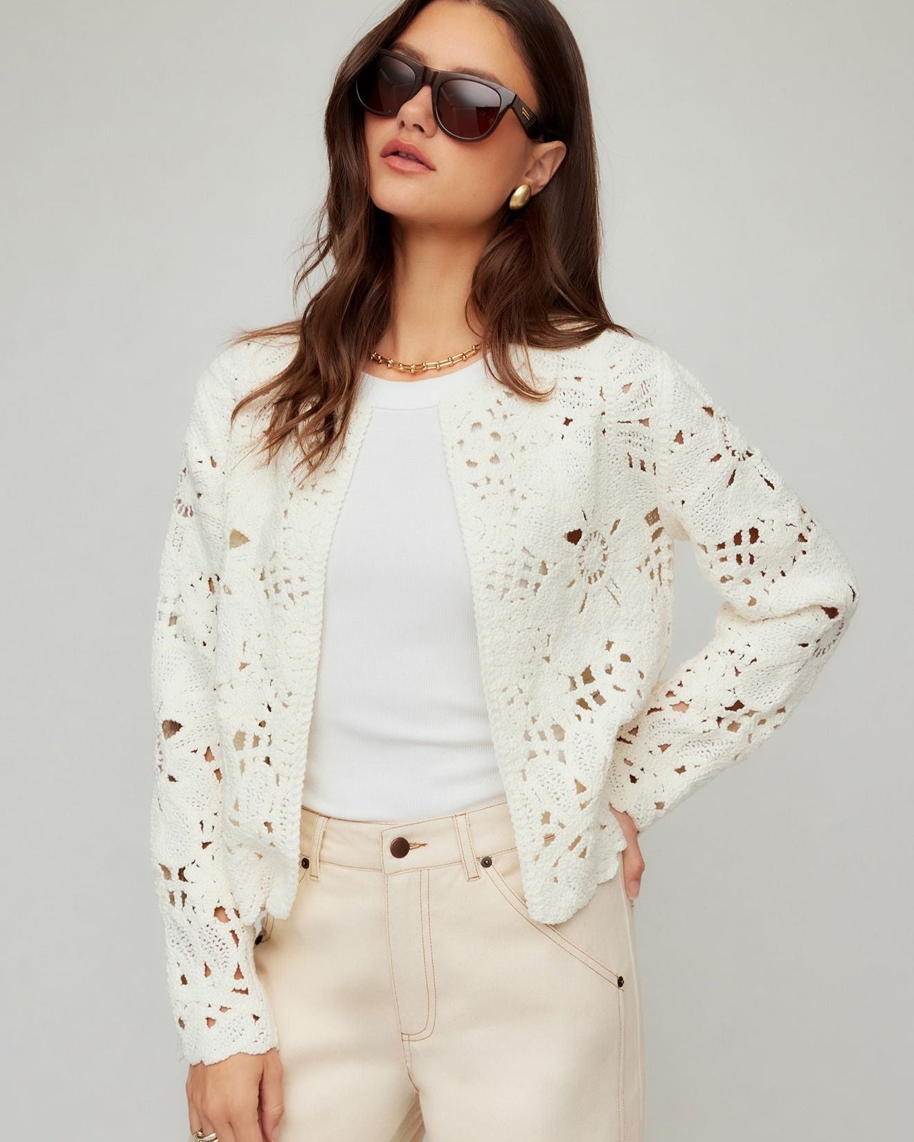 Woman wearing a white lace cardigan over a white shirt with beige pants on a gray background