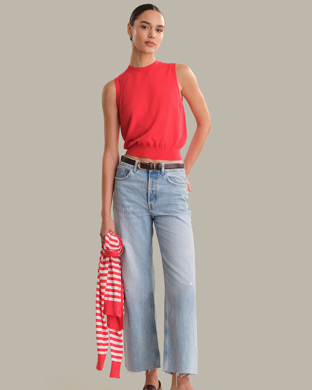 Woman wearing a red sleeveless top and blue jeans, holding a striped shirt, on a gray background