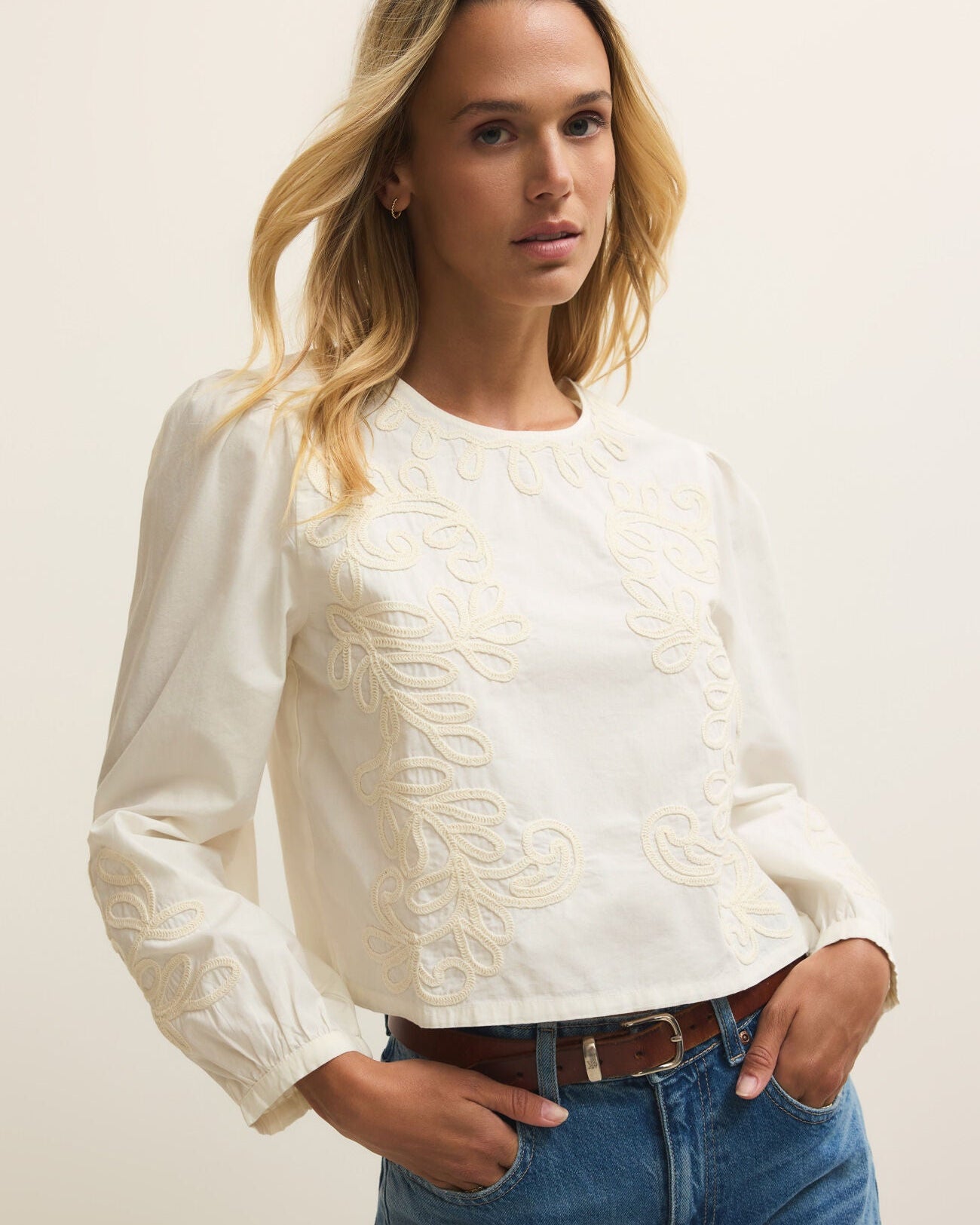 Woman wearing a white blouse with lace details and blue jeans against a plain background