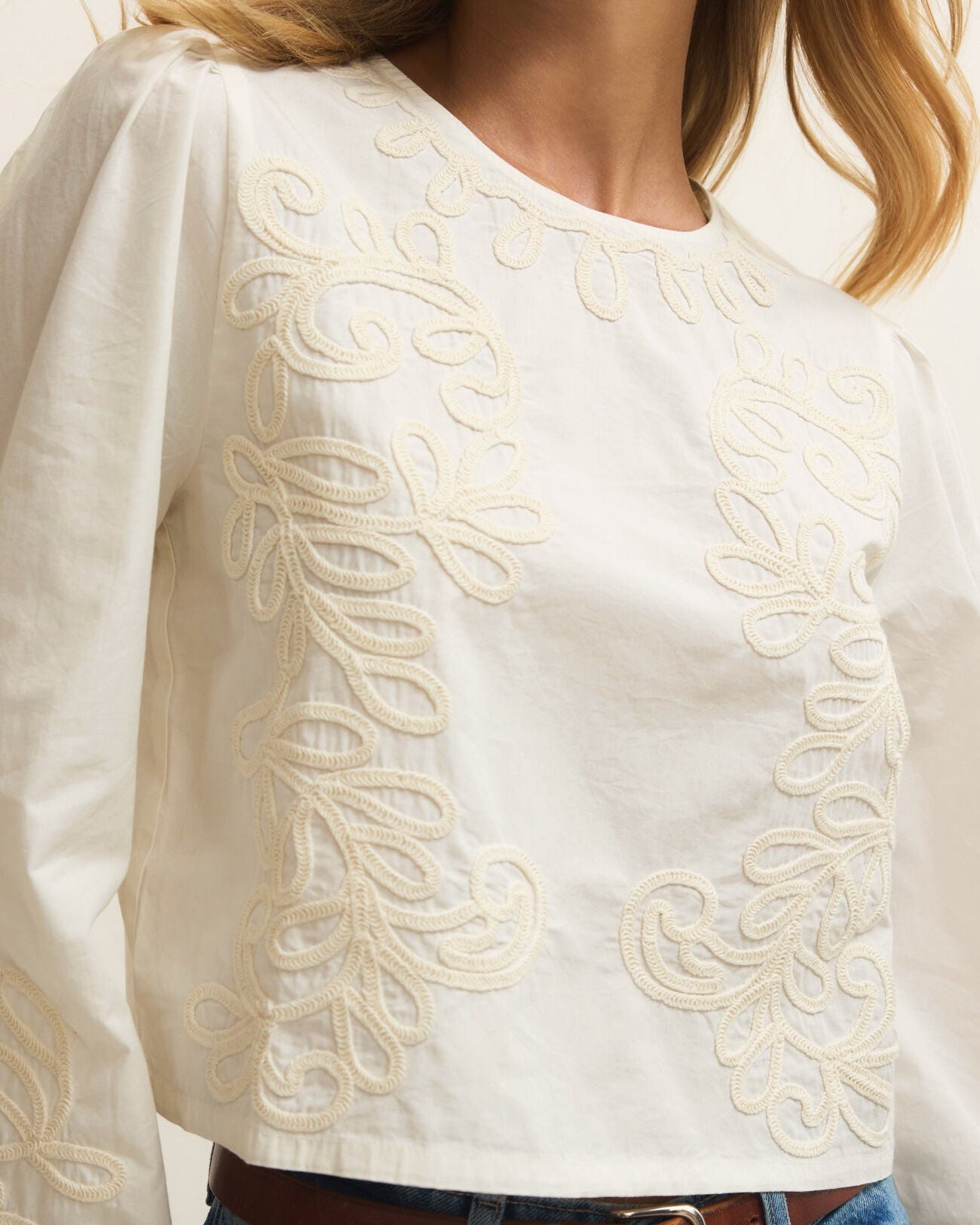 White blouse with intricate embroidery worn by a person, paired with blue jeans and a brown belt.