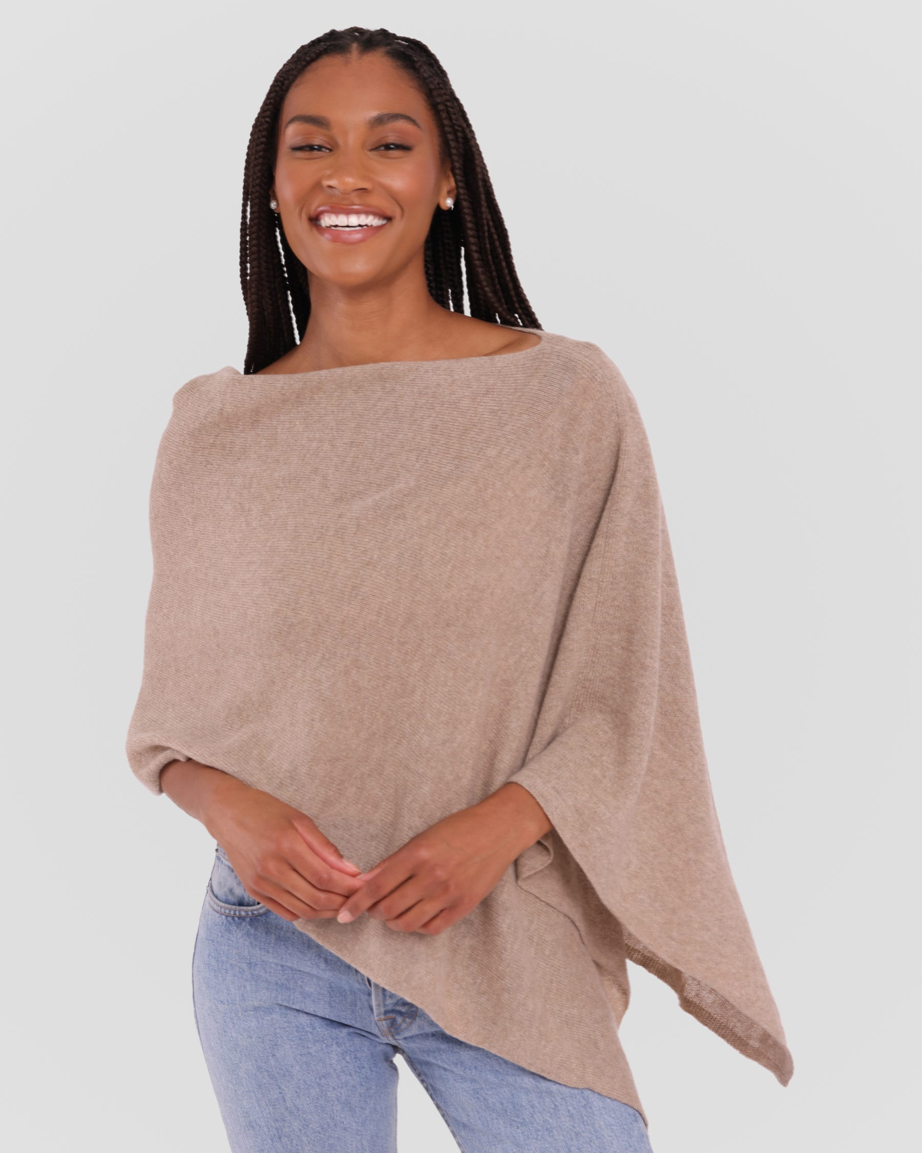 A woman wearing a light taupe knit poncho sweater and light blue jeans, standing against a solid light gray background.