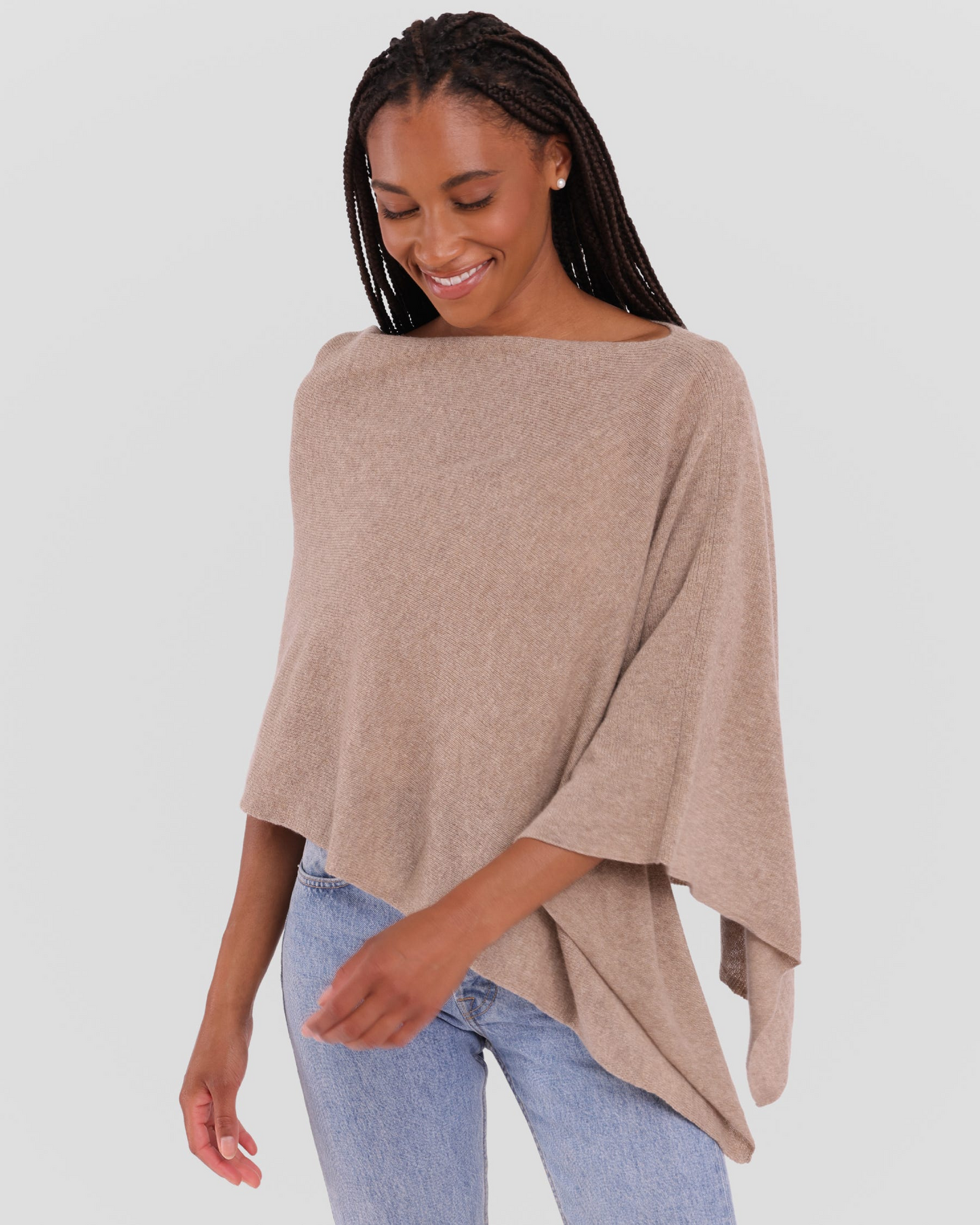 A woman wearing a light beige knit poncho top with a wide boat neckline and draped sleeves, paired with light blue jeans, against a solid light gray background.