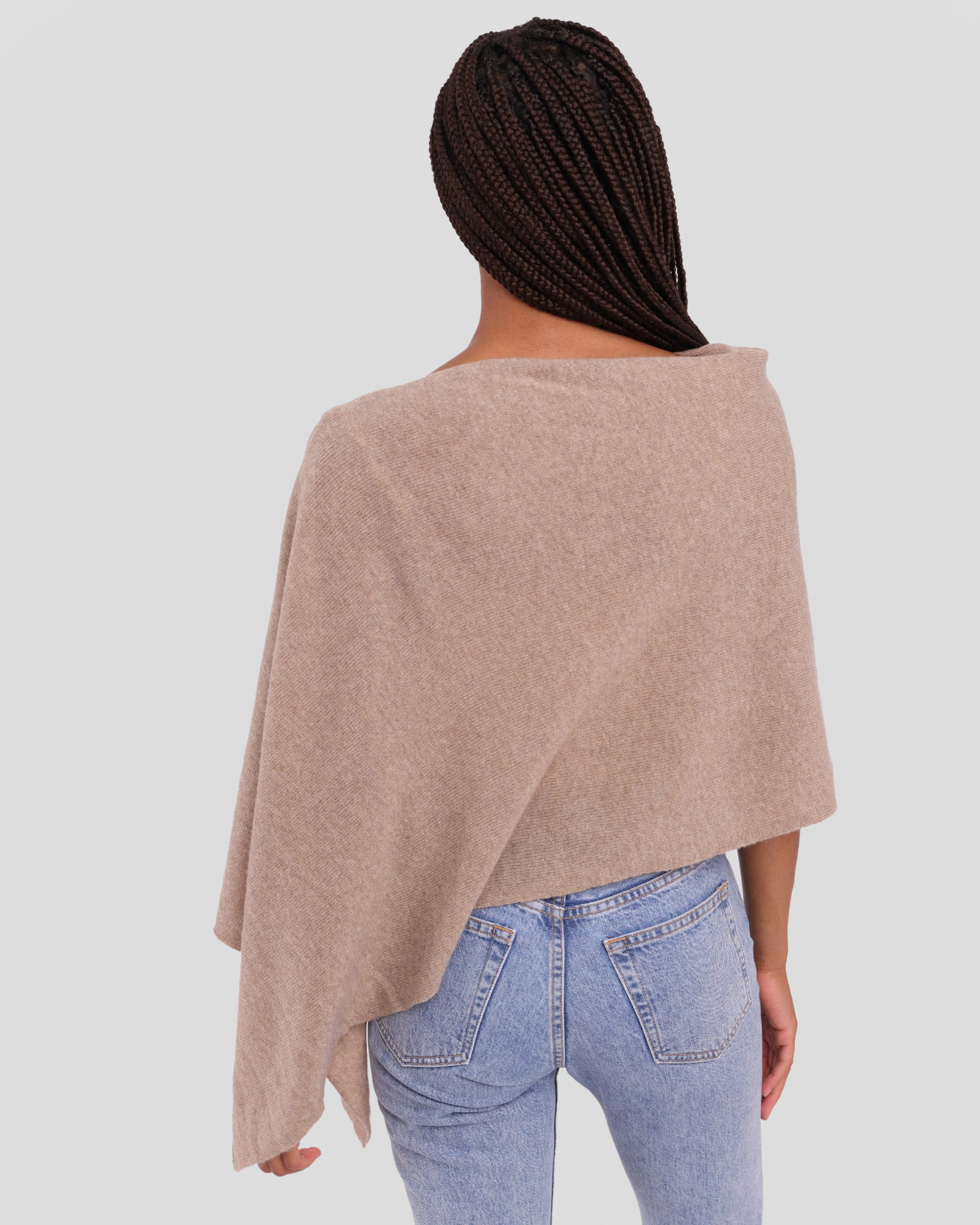 A woman viewed from behind wearing a light taupe knit poncho over light blue jeans, set against a solid light gray background.