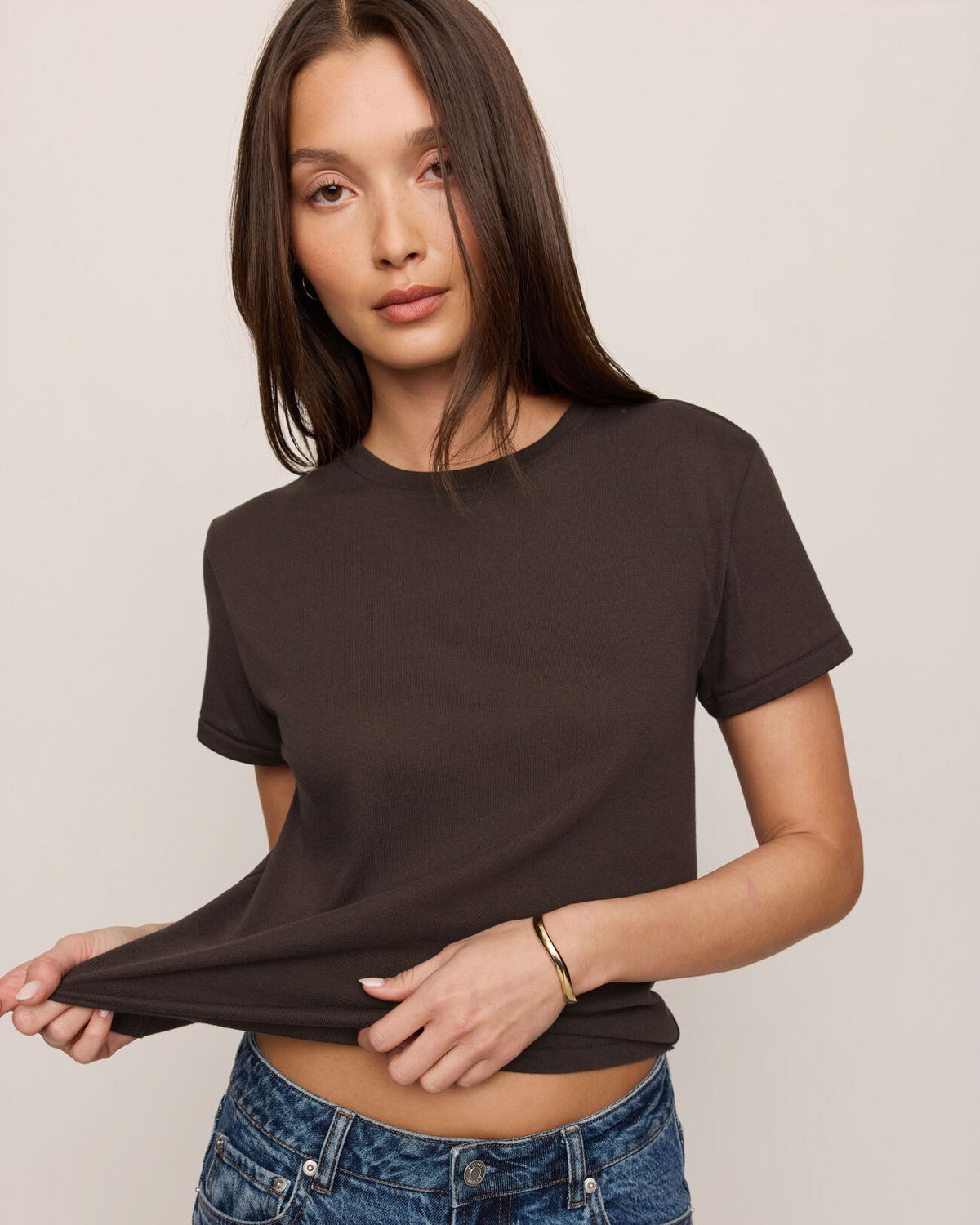 A woman wearing a dark brown short-sleeve crewneck t-shirt and blue jeans, lifting the hem of the shirt to show the fabric stretch, against a plain light beige background.