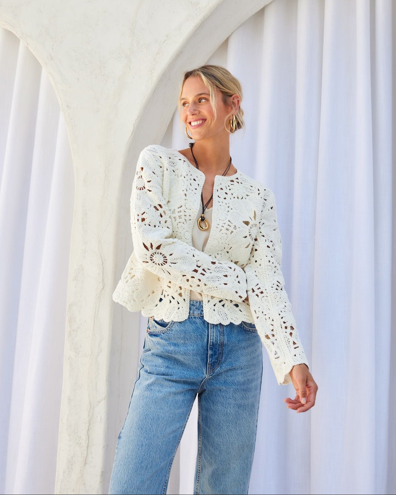 Woman wearing a white lace top and blue jeans standing in front of a white archway.