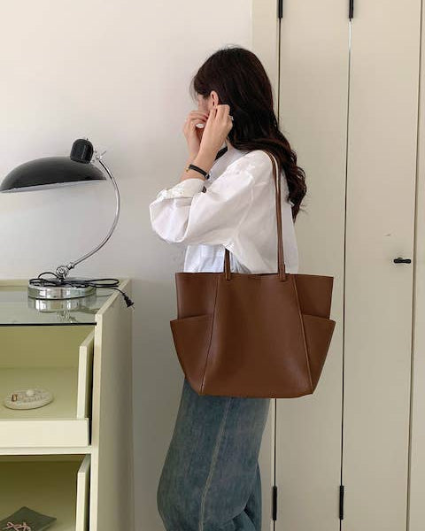 A woman wearing a white button-up shirt and blue jeans carries a large brown leather tote bag over her shoulder in a minimalist indoor setting.