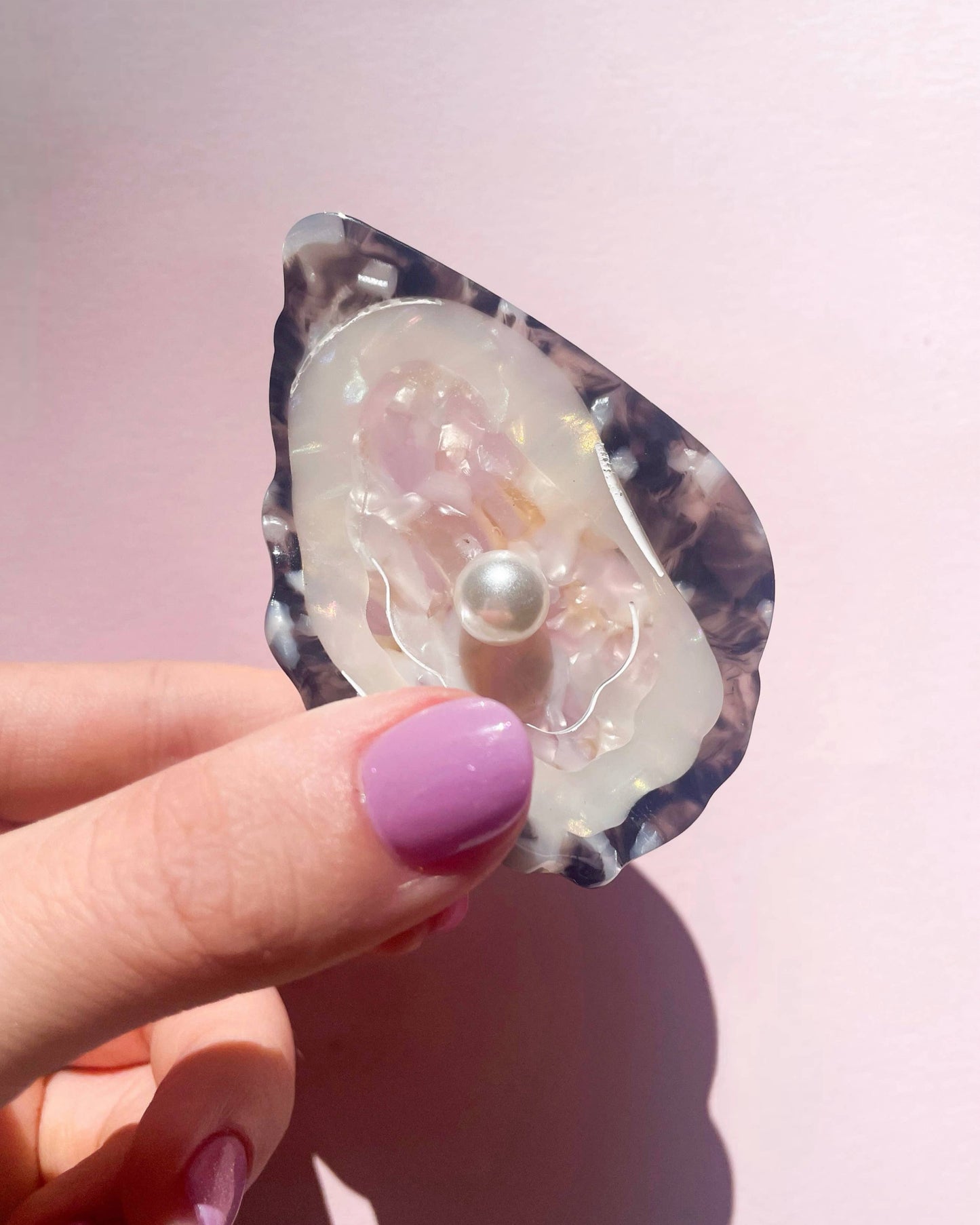 Oyster hair clip with a pearl inside, held by a hand with purple nail polish on a pink background