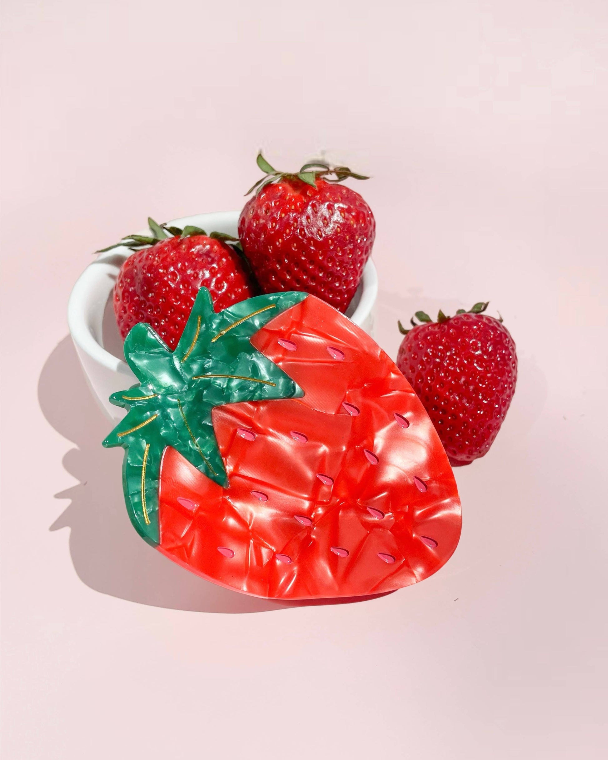 Strawberry-shaped dish with a red body and green leaves, surrounded by real strawberries on a pink background.