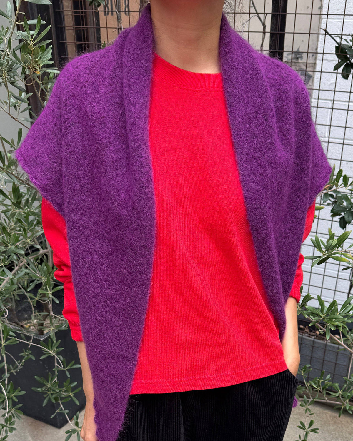A person wearing a bright purple fuzzy open-front shawl or cape over a vivid red long-sleeve top and black corduroy pants, standing outdoors near plants and a fence.