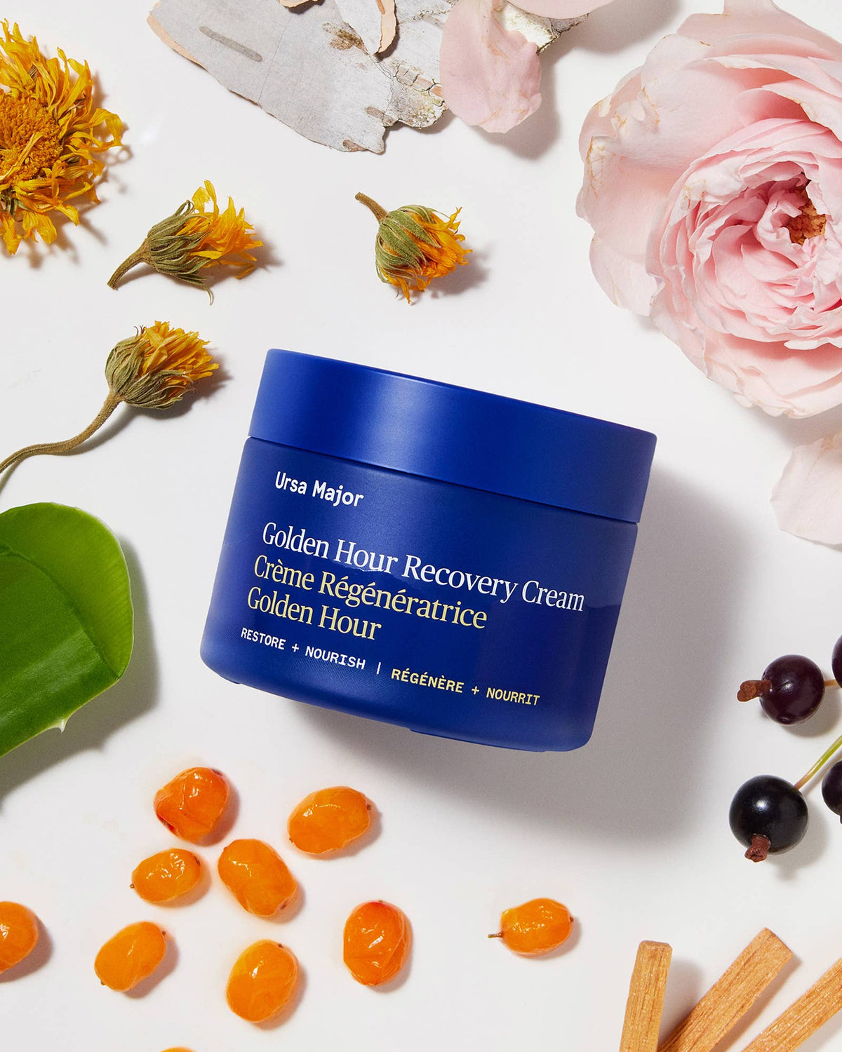 Blue jar of Ursa Major Golden Hour Recovery Cream surrounded by flowers and natural elements on a white background
