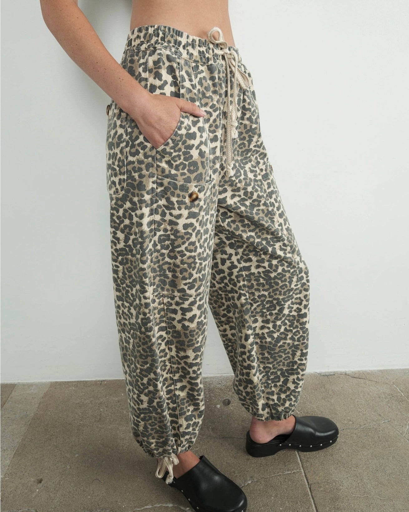 Side view of a model wearing the Aaron & Amber relaxed fit leopard pull on pant with hand in the pocket with black shoes against a white wall.