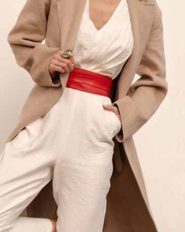 Person wearing a beige coat, white top, and white pants with a red belt against a neutral background
