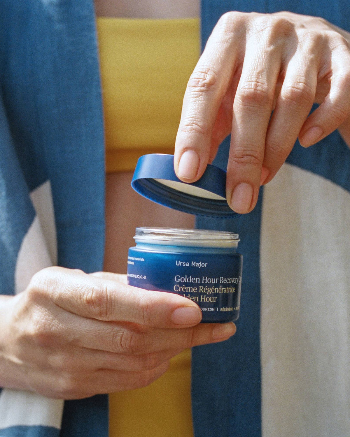 Person holding a blue jar of Ursa Major Golden Hour Renewal Cream with a blue towel in the background.