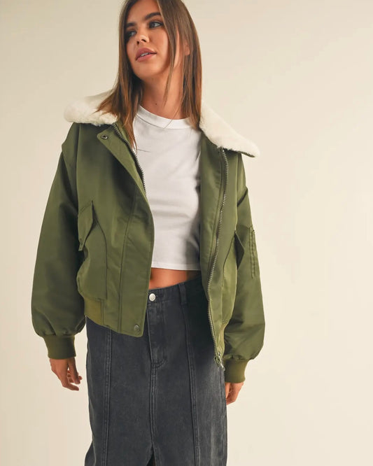 Image of  Fur Collared Bomber Jacket in Olive color wearing black skirt and white top on a beige background
