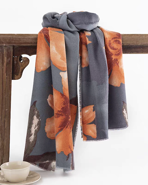 A gray scarf with a large orange floral pattern draped over a dark wooden table, with a small white teacup on a saucer in the lower left corner.